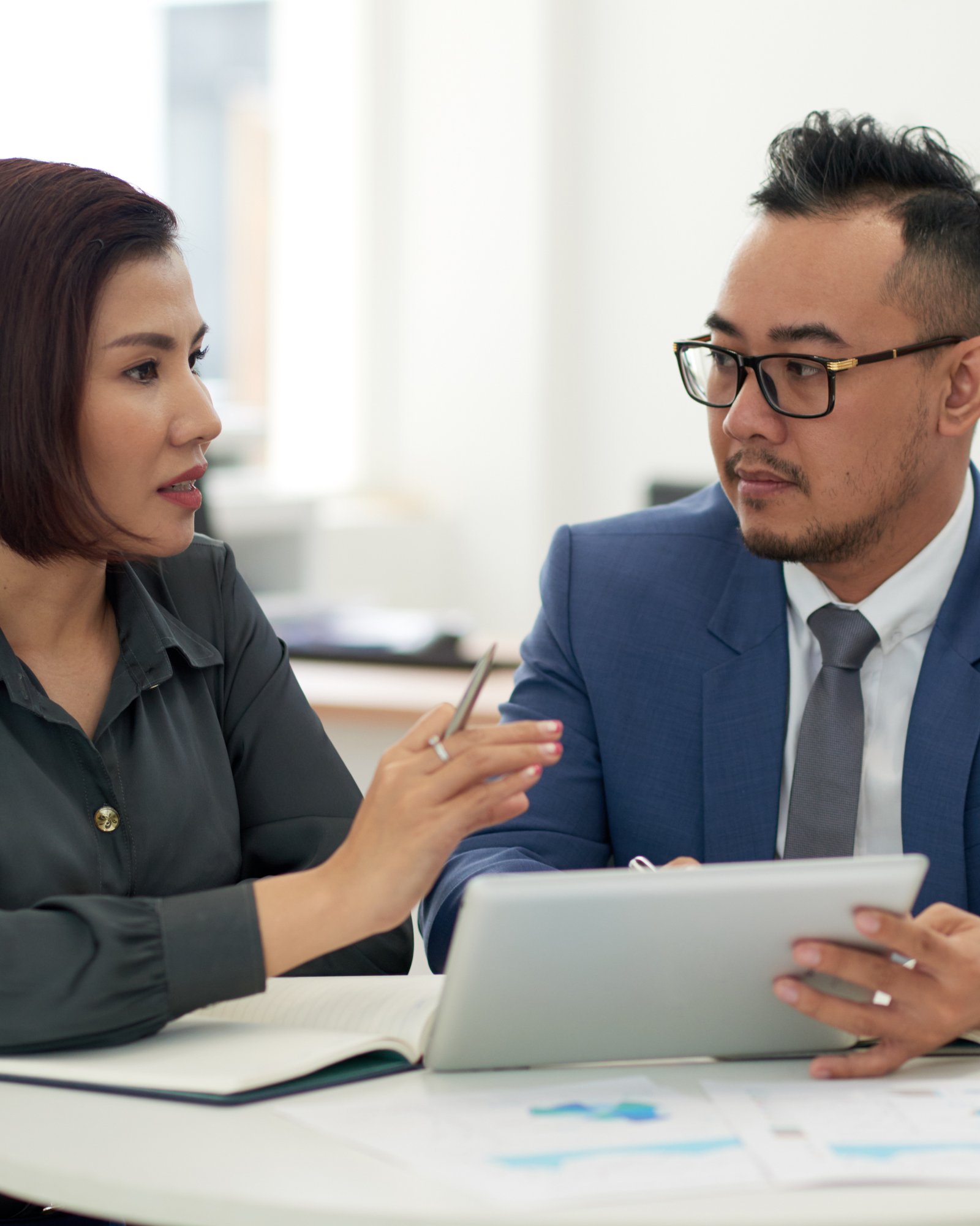 asian-man-woman-business-attire-sitting-indoors-with-tablet-talking